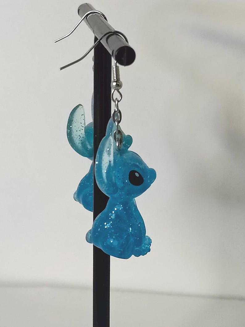 Pu&ograve; includere: Un paio di orecchini in resina glitterata blu con un design a personaggio dei cartoni animati. Gli orecchini hanno un gancio color argento e sono sospesi da un espositore in metallo nero. Il personaggio ha grandi occhi neri e una testa piccola.