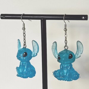 Pu&ograve; includere: Un paio di orecchini Stitch blu glitterati. Ogni orecchino presenta una piccola figura di Stitch seduta con grandi orecchie e occhi neri, sospesa a un gancio e una catena color argento. Gli orecchini sono esposti su un supporto nero su sfondo bianco.