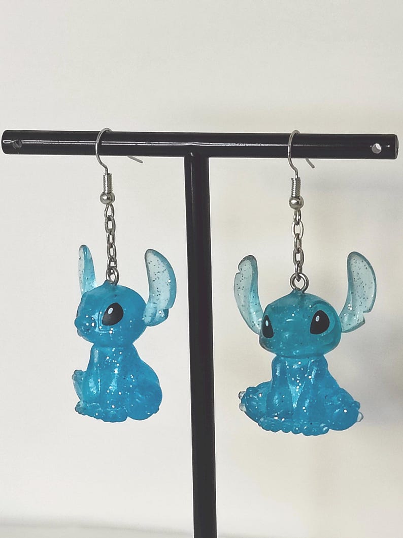 Pu&ograve; includere: Un paio di orecchini Stitch blu glitterati. Ogni orecchino presenta una piccola figura di Stitch seduta con grandi orecchie e occhi neri, sospesa a un gancio e una catena color argento. Gli orecchini sono esposti su un supporto nero su sfondo bianco.