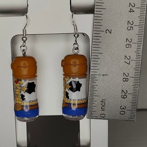 May include: A pair of dangle earrings featuring clear plastic bottles with a brown lid and a printed image of Woody from Toy Story. The image shows Woody's cowboy hat, vest, and jeans. The bottles are filled with clear glitter.