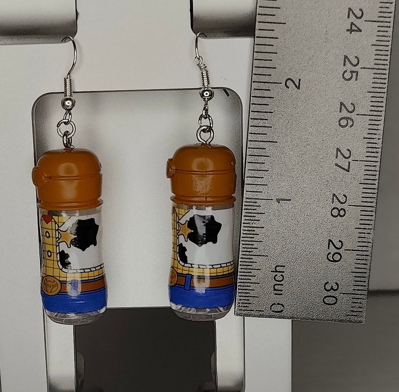 May include: A pair of dangle earrings featuring clear plastic bottles with a brown lid and a printed image of Woody from Toy Story. The image shows Woody's cowboy hat, vest, and jeans. The bottles are filled with clear glitter.
