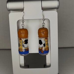 May include: A pair of dangle earrings featuring a clear plastic bottle with a brown lid and a printed image of Woody from Toy Story. The bottle is filled with a clear liquid and has a silver hook.