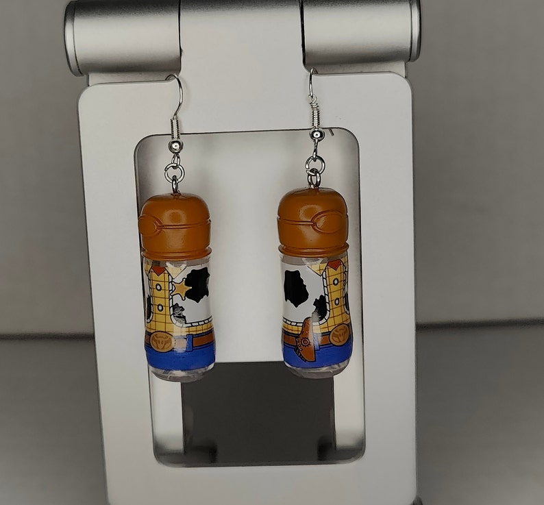 May include: A pair of dangle earrings featuring a clear plastic bottle with a brown lid and a printed image of Woody from Toy Story. The bottle is filled with a clear liquid and has a silver hook.