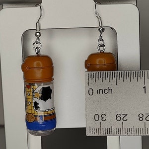 May include: A pair of earrings featuring small brown plastic bottles with a cartoon image of Woody from Toy Story. The bottles are approximately 1 inch tall.