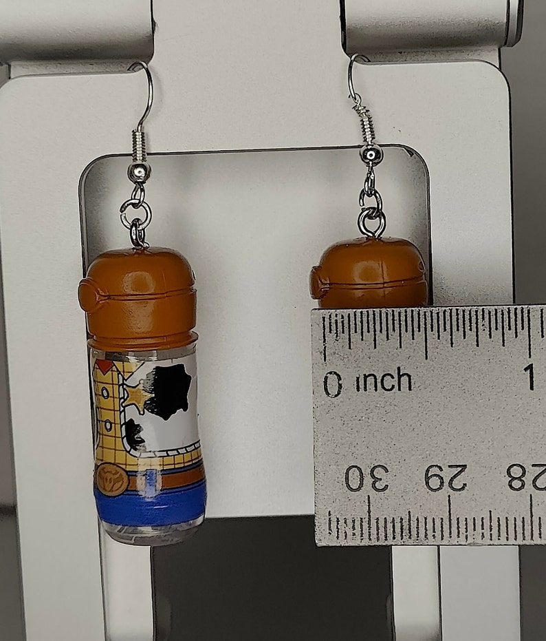 May include: A pair of earrings featuring small brown plastic bottles with a cartoon image of Woody from Toy Story. The bottles are approximately 1 inch tall.