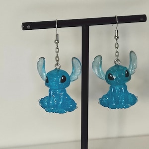 Pu&ograve; includere: Un paio di orecchini blu di Stitch. Ogni orecchino presenta una piccola figura di Stitch blu glitterata con grandi orecchie e occhi neri, sospesa a una catena e un gancio color argento. Gli orecchini sono esposti su un supporto nero.