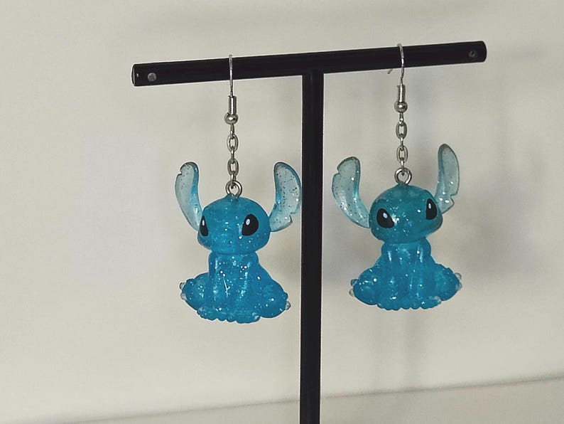 Pu&ograve; includere: Un paio di orecchini blu di Stitch. Ogni orecchino presenta una piccola figura di Stitch blu glitterata con grandi orecchie e occhi neri, sospesa a una catena e un gancio color argento. Gli orecchini sono esposti su un supporto nero.