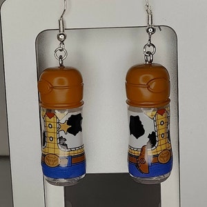 May include: A pair of dangle earrings featuring a brown plastic bottle with a cartoon image of Woody from Toy Story. The bottle is filled with a clear liquid and has a blue and yellow design.