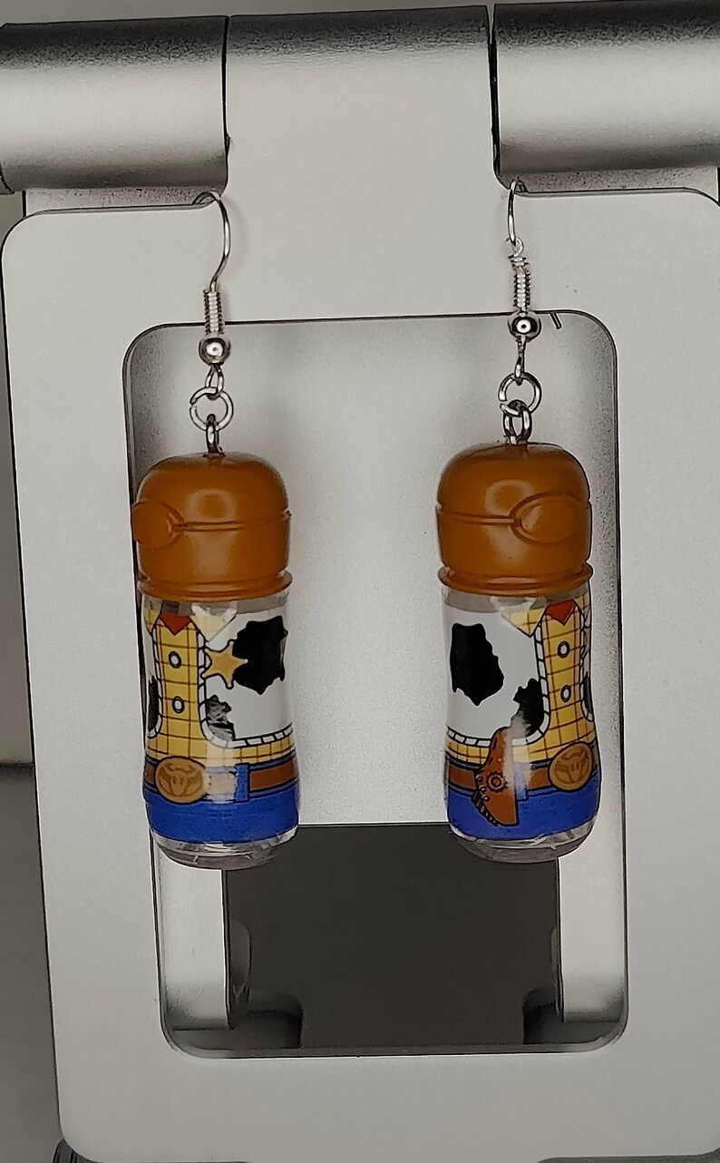 May include: A pair of dangle earrings featuring a brown plastic bottle with a cartoon image of Woody from Toy Story. The bottle is filled with a clear liquid and has a blue and yellow design.