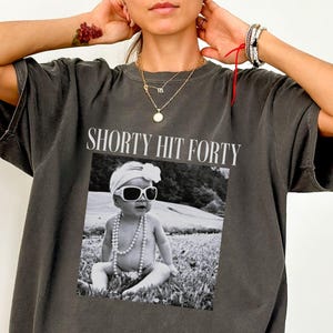 Peut inclure: T-shirt gris foncé avec l'inscription "SHORTY HIT FORTY" au-dessus d'une photo en noir et blanc d'un bébé portant des lunettes de soleil et un collier de perles. Le mot "Pepper" est imprimé en bas.