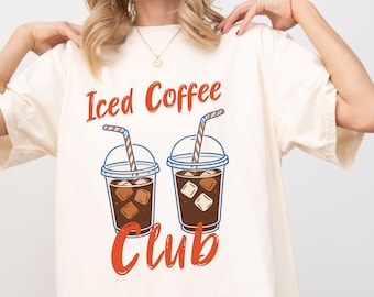 以下が含まれることがあります： アイボリー色のTシャツ。ストロー付きのアイスコーヒー2杯のグラフィック。赤字で「Iced Coffee Club」とプリントされています。ゆったりとしたフィット感のシャツです。