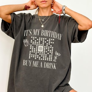 May include: Dark gray t-shirt with the text "IT'S MY BIRTHDAY BUY ME A DRINK" in white. The shirt features a QR code design with cocktail and gift illustrations. The shirt is a relaxed fit.