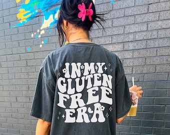 May include: A person wearing a black t-shirt with the text "In My Gluten Free Era" in white. The shirt has a retro font style and is decorated with small white flowers.