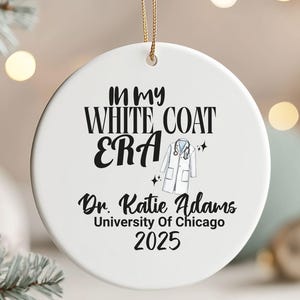 May include: White ceramic Christmas ornament with gold string. The ornament features the text "IN MY WHITE COAT ERA" and "Dr. Katie Adams University Of Chicago 2025". A graphic of a white coat and stethoscope is also included.