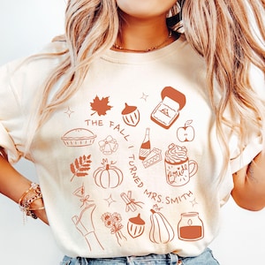 May include: Ivory-colored t-shirt with a fall-themed graphic in brown. The design includes a leaf, pie, pumpkin, ring box, apple, and a mug that says "Bride". Text on the shirt reads "The Fall I Turned Mrs. Smith."