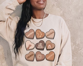 May include: A beige sweatshirt with a graphic print of hearts featuring the words "Educated", "Melanin", "I Am Black History", and "Elevated".