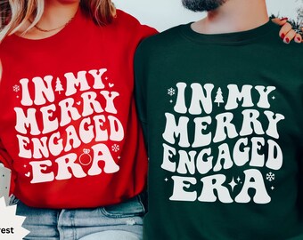 May include: Red and forest green sweatshirts with the text "IN MY MERRY ENGAGED ERA" in white. The design includes snowflakes, a Christmas tree, hearts, and a ring. The sweatshirts are a festive choice for couples.