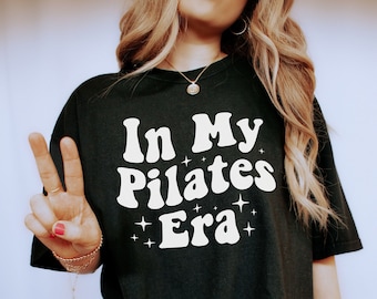 Pode incluir: Uma camiseta preta oversized com o texto "In My Pilates Era" em branco com uma fonte retrô e estrelas brilhantes. A modelo está usando shorts de estampa de leopardo e tênis brancos.