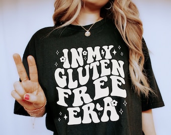 May include: A black t-shirt with white text that reads "In My Gluten Free Era" in a retro font. The shirt has a flower graphic and stars around the text. The person wearing the shirt is making a peace sign with their hand.