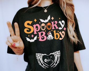 May include: A black t-shirt with the text "Spooky Baby" in colorful letters. The shirt features a ghost, spider, and bats. A pair of skeleton hands are holding a heart with a skeleton baby inside.