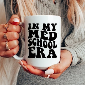 以下が含まれることがあります： レトロなフォントで「In My Med School Era」と書かれた黒いテキストが施された白いセラミックマグカップです。