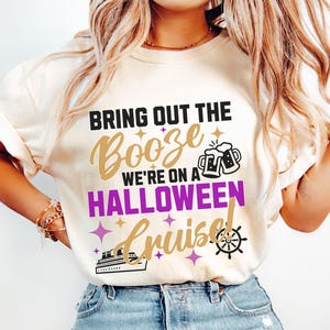 May include: Ivory-colored t-shirt with the text "Bring Out The Booze We're On A Halloween Cruise!" in black, gold, and purple. The design includes beer mugs, stars, a cruise ship, and a ship's wheel.