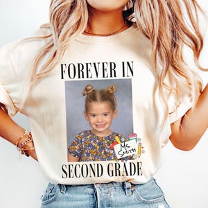 Może przedstawiać: Kremowa koszulka z napisem "FOREVER IN SECOND GRADE" nad zdjęciem szkolnym małej dziewczynki. Koszulka zawiera również imię "Ms. Smith" i grafikę ołówka.