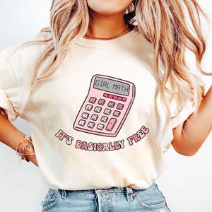 Puede incluir: Camiseta color marfil con un diseño de calculadora rosa que muestra "GIRL MATH". Debajo, la frase "IT'S BASICALLY FREE" está impresa en una fuente curva. La palabra "Ivory" está impresa en la parte inferior.