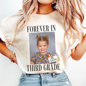 May include: An ivory short-sleeve t-shirt featuring a central graphic with the text "FOREVER IN THIRD GRADE" and a photograph of a young child with hair in two buns. The graphic includes a small white sign with the handwritten text "Ms. SMITH".