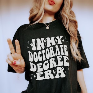 May include: A black t-shirt with a white graphic that says "In My Doctorate Degree Era" with a flower and lightning bolt design.