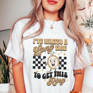 Puede incluir: Camiseta blanca con un gráfico dorado de una esfera de reloj con un anillo de diamantes en él. El texto "He esperado mucho tiempo para conseguir este anillo" está escrito en una fuente retro.