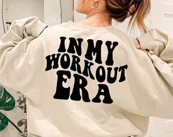 May include: A beige sweatshirt with the text "IN MY WORKOUT ERA" printed in black retro-style lettering on the back.