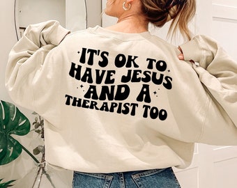 May include: A beige sweatshirt with the text "It's ok to have Jesus and a therapist too" printed on the back in black.
