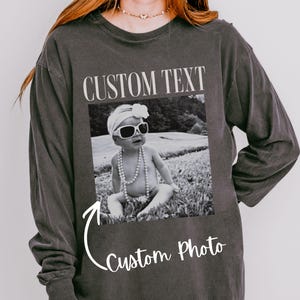May include: Dark gray long-sleeve shirt with a custom photo design. The design features a black and white photo of a baby wearing sunglasses and a pearl necklace. The text "CUSTOM TEXT" is above the photo, and "Custom Photo" is below.