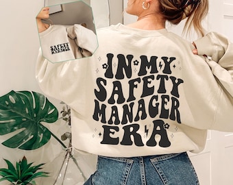 May include: A cream-colored sweatshirt with the text "In My Safety Manager Era" in black retro-style lettering. The text is surrounded by small black stars and flowers.