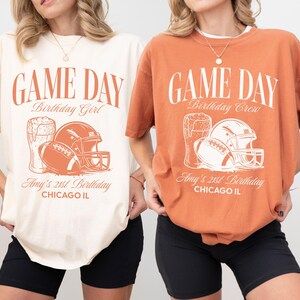 Op de afbeelding: Twee T-shirts, &eacute;&eacute;n ivoorkleurig en &eacute;&eacute;n yam-kleurig, met een voetbalthema. Elk shirt heeft de tekst "GAME DAY" en een afbeelding van een voetbal, helm en bier. De tekst "Birthday Girl" en "Birthday Crew" worden ook weergegeven.