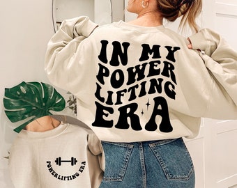 May include: A beige sweatshirt with the text "IN MY POWERLIFTING ERA" in black, retro-style lettering on the back. The front of the sweatshirt has a smaller version of the text with a dumbbell graphic.