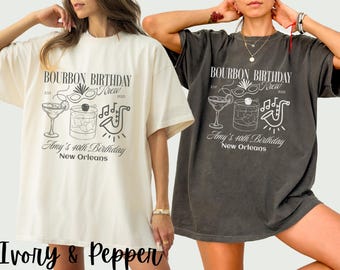 May include: Two oversized t-shirts, one ivory and one pepper, featuring a graphic design. The design includes text that reads "BOURBON BIRTHDAY Krew" and "Amy's 40th Birthday New Orleans" with cocktail and music-themed illustrations.