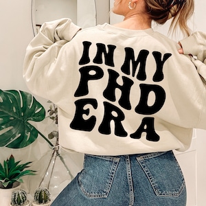 May include: A cream-colored sweatshirt with the text "In My PhD Era" printed in black, retro-style lettering.