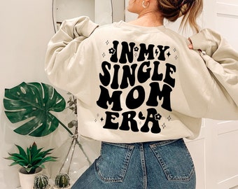 May include: A beige sweatshirt with the text "In My Single Mom Era" in black retro-style lettering. The text is surrounded by small black stars and flowers.