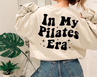 May include: A beige sweatshirt with the text "In My Pilates Era" in black, retro-style lettering with sparkling stars.