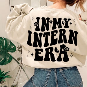 May include: A cream-colored sweatshirt with the text "IN MY INTERN ERA" in black retro-style lettering. The text is surrounded by small black stars and flowers.