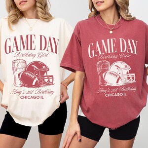 Op de afbeelding: Twee T-shirts met korte mouwen, &eacute;&eacute;n ivoorkleurig en &eacute;&eacute;n baksteenkleurig, met een voetbalthema. Het ontwerp bevat een voetbal, helm en bierpul, met de tekst "GAME DAY" en "Amy's 21st Birthday, Chicago IL."