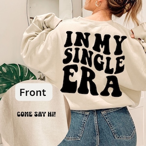 Może przedstawiać: Beżowa bluza z nadrukiem "IN MY SINGLE ERA" w czarnym, retro stylu na plecach. Z przodu bluza ma napis "COME SAY HI!" w kolorze czarnym.