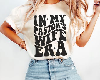 以下が含まれることがあります： 黒いレトロな文字で「In My Pastor's Wife Era」とプリントされた白いTシャツです。