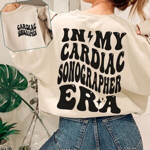 Puede incluir: Una sudadera de color crema con el texto "In My Cardiac Sonographer Era" en letras negras de estilo retro. El texto está en la parte posterior de la sudadera.