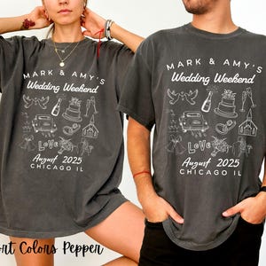 Pode incluir: Duas t-shirts cinzas escuras com texto branco e ilustrações. O texto nas t-shirts diz "Mark & Amy's Wedding Weekend" com a data "August 2025" e a localização "Chicago IL". As t-shirts apresentam ilustrações de ícones relacionados com o casamento, como pombos, anéis, uma igreja, um bolo e um carro.