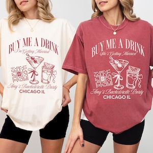 Peut inclure: Deux femmes portant des t-shirts avec le texte "Buy Me A Drink" en rouge. Le texte est sur un t-shirt blanc et un t-shirt rouge. Les t-shirts ont également un graphique d'un verre à cocktail et d'une petite bouteille. Le texte sous le graphique dit "Amy's Bachelorette Party Chicago IL".