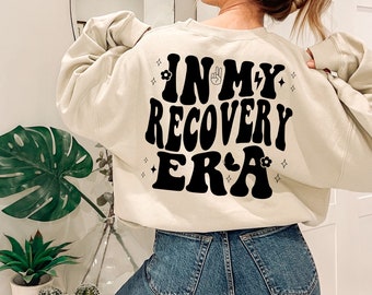 以下が含まれることがあります： 薄いベージュのトレーナーで、背面に「IN MY RECOVERY ERA」という黒いグラフィックが描かれており、ピースサインと花が描かれています。