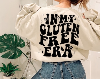 May include: A beige sweatshirt with the text "IN MY GLUTEN FREE ERA" printed in black retro-style lettering. The text is surrounded by small black stars and flowers.
